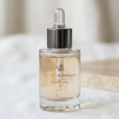 A photorealistic product shot of a clear glass serum bottle filled with a light amber liquid. The front features a delicate gold logo of a flower with text that reads "ÉCLAT | BOTANIQUE" and "SERUM VITAL" below it. At the base, small text states "30ml e 1.0 fl. oz. | PARIS | London". The bottle has a shiny metallic silver collar and a clear glass dropper cap, standing on a white linen surface with a blurred background.