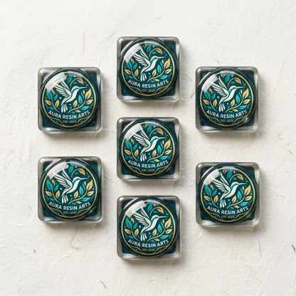Seven square resin stickers arranged in a diamond pattern on a white textured background, each featuring the Aura Resin Arts logo — a white hummingbird in flight surrounded by teal and gold foliage inside a circular gold border, with “Aura Resin Arts Est. 2023” text on a dark teal background.