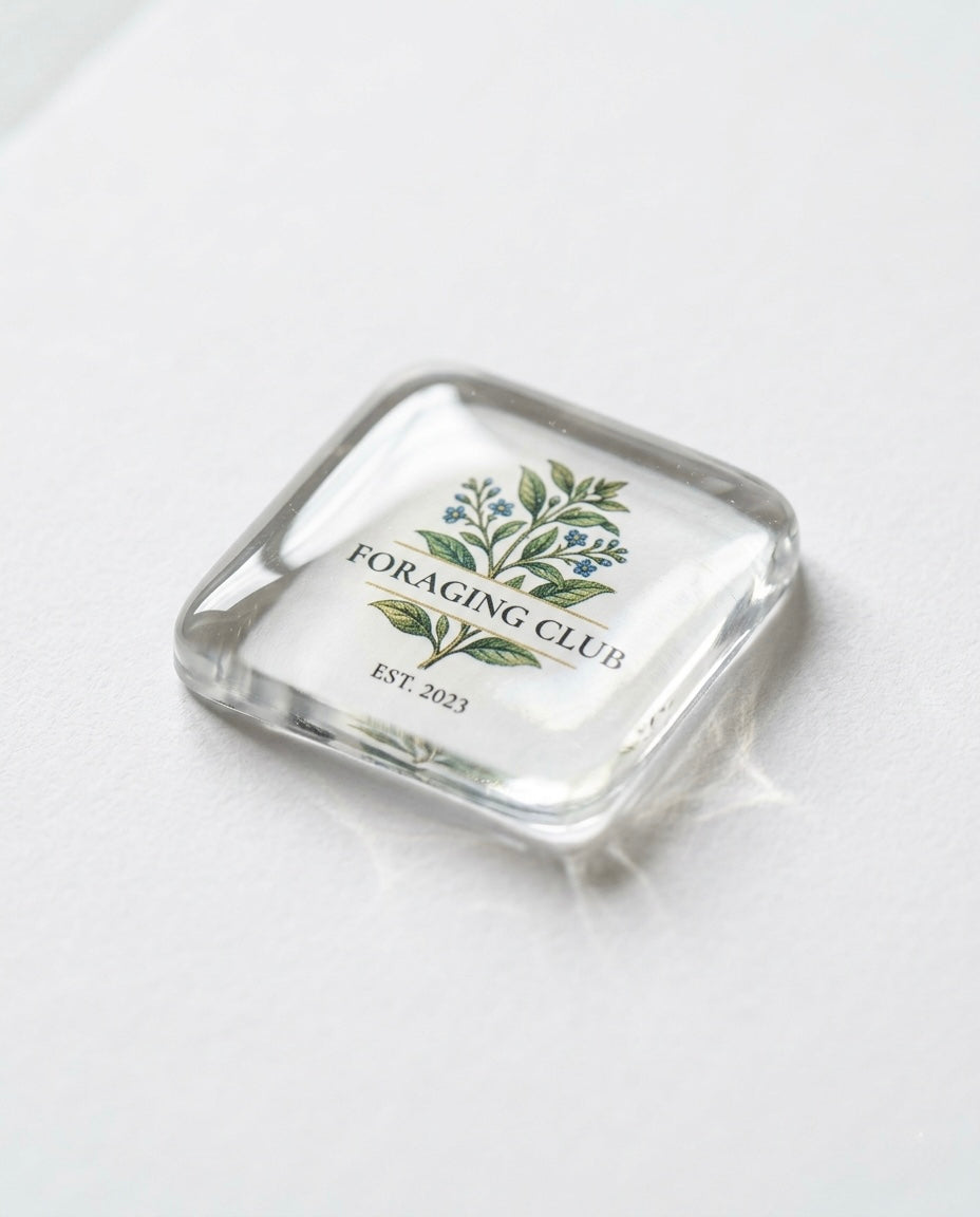 A single clear square resin sticker photographed at an angle on a white surface, featuring a botanical illustration of green leaves and small blue flowers above the text “Foraging Club Est. 2023” in elegant serif typography on a white background.
